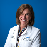 Dr. Beth Karlan smiling and wearing her UCLA lab coat with a blue backdrop behind.