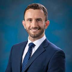 Smiling man in navy suit and tie against blue studio background.
