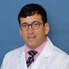 A person wearing a white lab coat, pink shirt, and striped tie stands against a blue background.