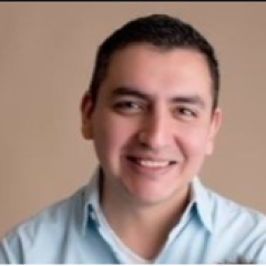A man with short brown hair, wearing a blue collared shirt, smiling in front of a beige background