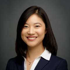A woman with black hair, wearing a navy suit jacket with a white shirt and necklace