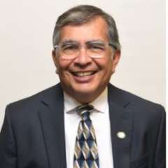 A man with short grey hair, wearing a suit and tie with glasses, smiling in front of a blank background