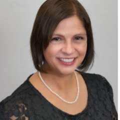 A woman with straight brown hair, wearing a black dress with a necklace, smiling in front of a blank background
