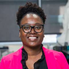 Rhonda Souvenir wearing black sunglasses, black ruffle blouse, and pink blazer. Background is blurry.