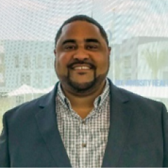A man with short black hair and a goatee, wearing a blazer and collared shirt, smiling in front of a building window