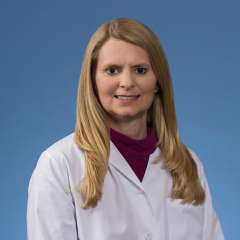 Blonde woman, Kailee Gaffny, wearing a white lab coat over a maroon top, stands in a professional, confident pose against a blue background.