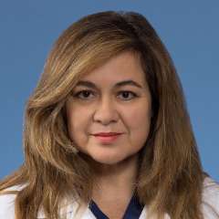Professional headshot Ana Valenzuela with long, brown, layered hair, wearing a white medical coat over blue scrubs with a calm, confident expression.