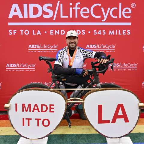Thomas Johansen stands behind his bicycle in front of an AIDS/LifeCycle sign at the end of the ride in Santa Monica