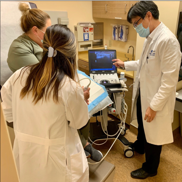 A man with dark hair points at an ultrasound machine while teaching two women. All three are focused on the screen, and the setting appears to be a medical or clinical environment.